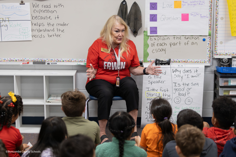 Ms. Plemmons sits in front of a classroom in a chair, as third graders site on the floor listening to her speak
