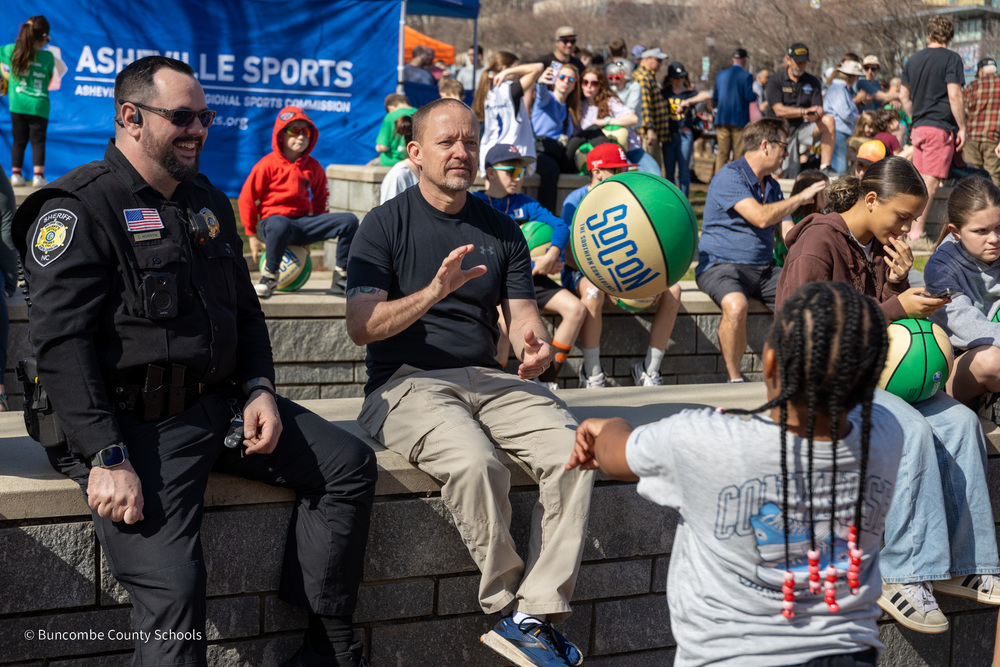 A student passes a basketball to an officer sitting on a wall in Pack Square Park. Another deputy sitting on the wall smiles.