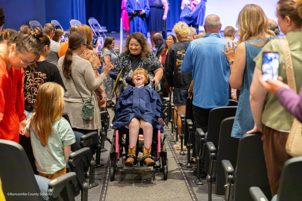 Student being rolled in wheelchair while crowd smiles and claps