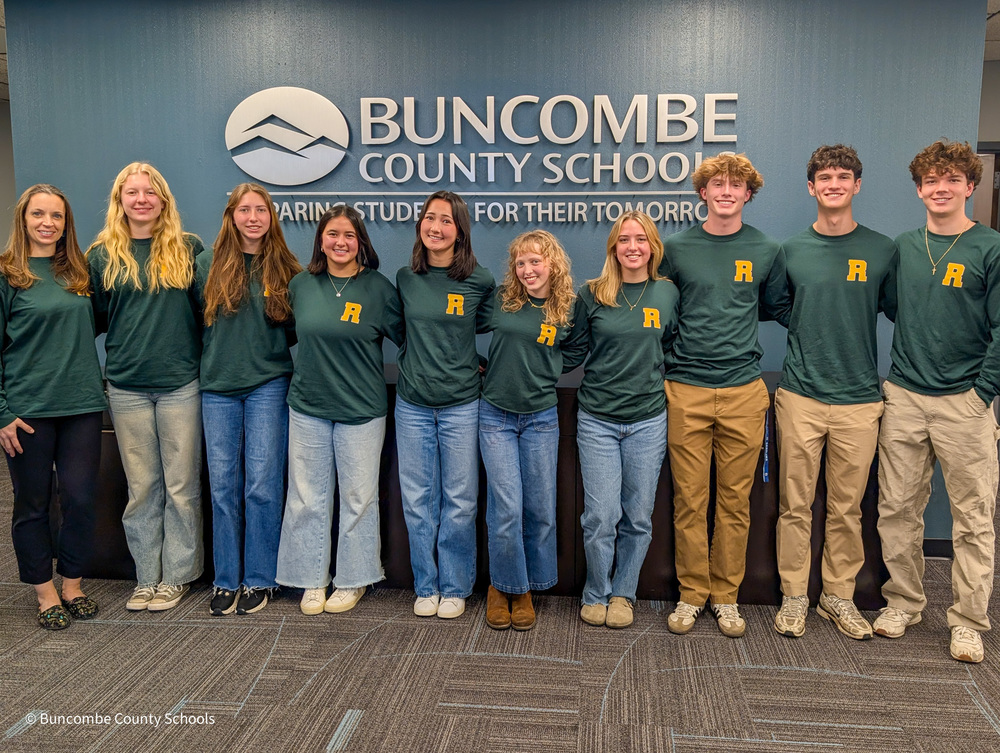 Ms. Brouwer and her Environmental Club students stand in a row posing for a photo in front of the Buncombe County Schools sign in the superintendent's lobby.
