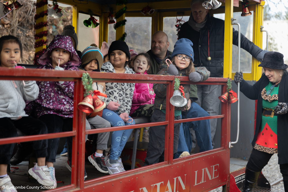 Group of diverse students riding on the train car and looking over at Santa and Mrs. Claus waving at them. 