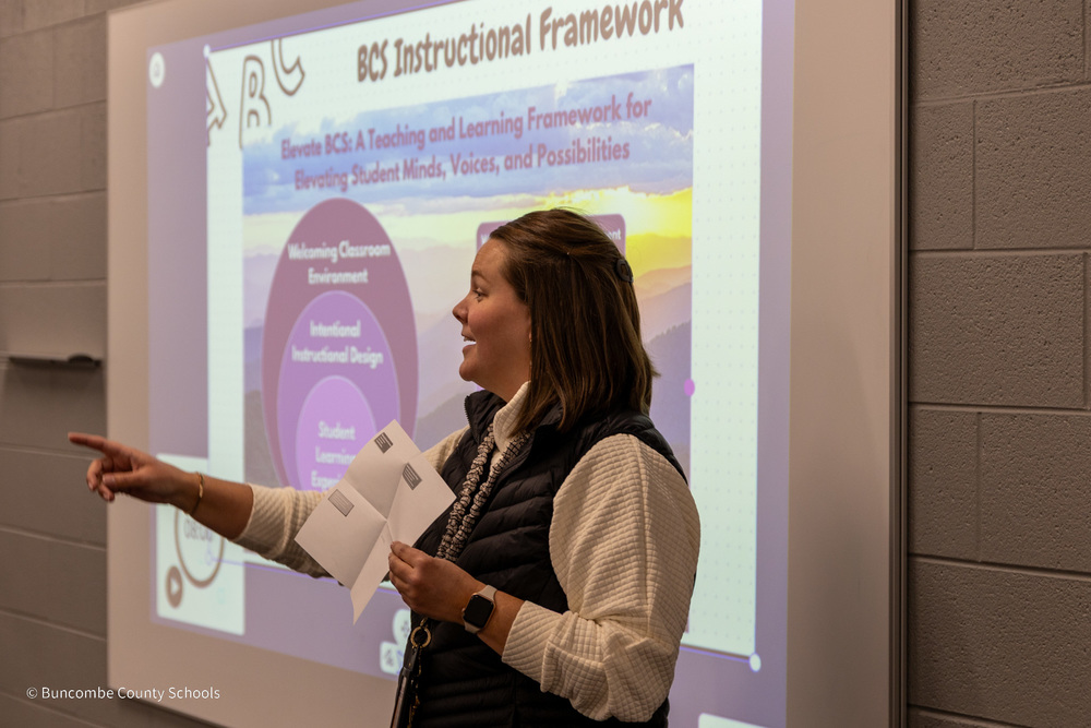 Martha Tallent stands in front of smartboard projecting a graphic of the Elevate BCS instructional Framework. She is smiling and pointing.