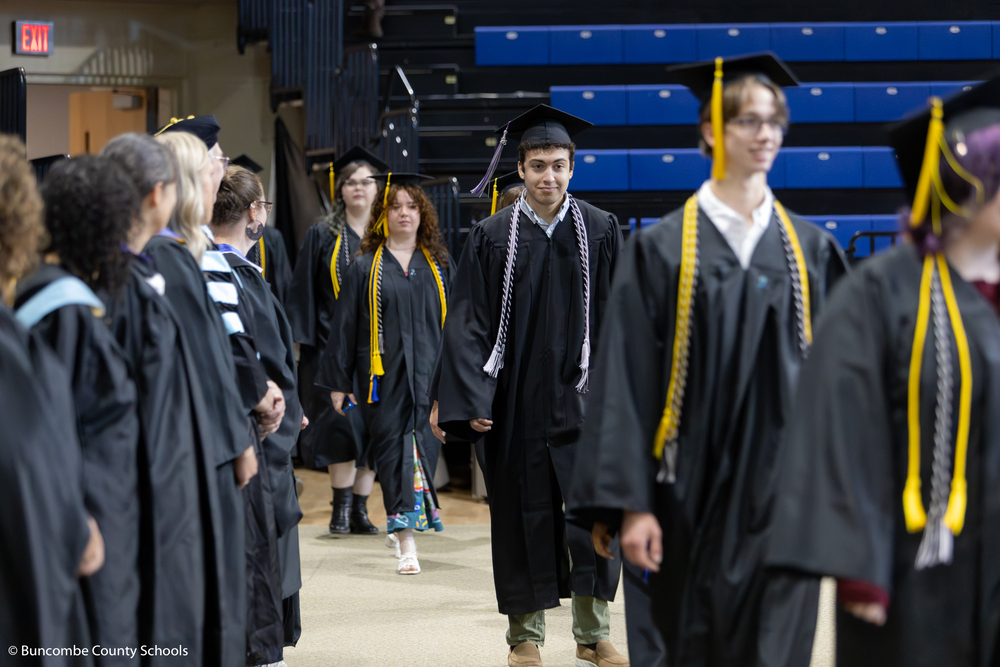 A line of students dressed in black caps and gowns walks in a line into the arena.