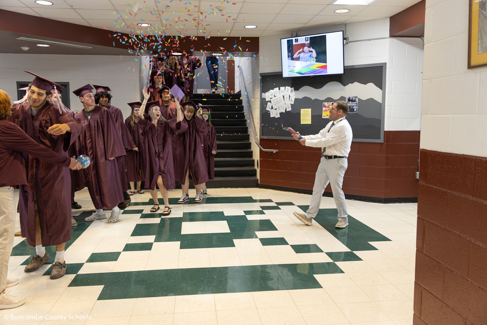 A man shoots a confetti cannon at Owen seniors dressed in maroon caps and gowns walking down the steps.