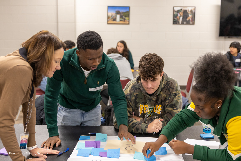 Dr. Johnson leans on a table surrounded by three students who are placing post-it notes on chart parper