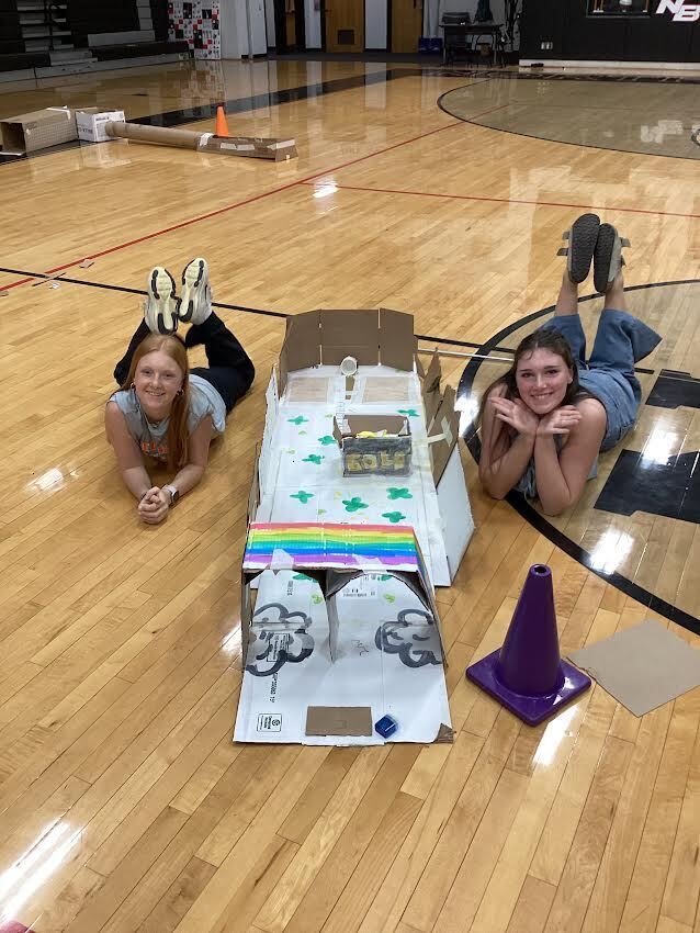 Two girls laying on the gym floor next to a putt-putt course they made.