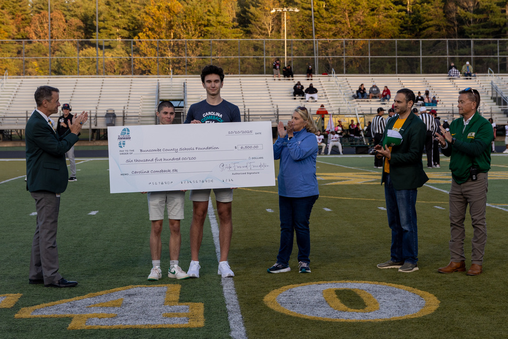 BCS Superintendent Dr. Rob Jackson, Pablo Bermudez, Matthew Balaban, Christy Cheek, Principal Ben Alexander, and Assistant Principal Steve McCurry stand on the 40 yard line of the A.C. Reynolds football field. Pablo and Matthew are holding a giant novelty check for $6,500 made out to the Buncombe County Schools Foundation.