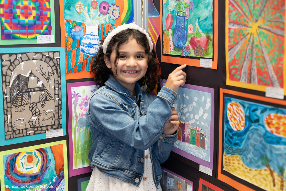 First grader smiling at the camera and pointing to her artwork. 