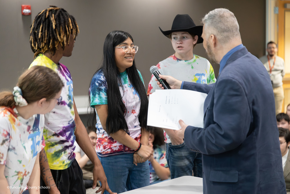 Four students in matching tie-dye T-shirts stand together at an event as one student speaks into a microphone held by Buncombe County Schools Chief Communications Officer Ken Ulmer.
