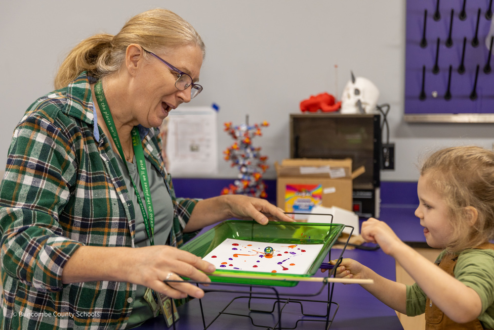 A teacher holds a tray with a metal ball and paint. A student is controlling the ball with a magnet beneath the tray to create art.