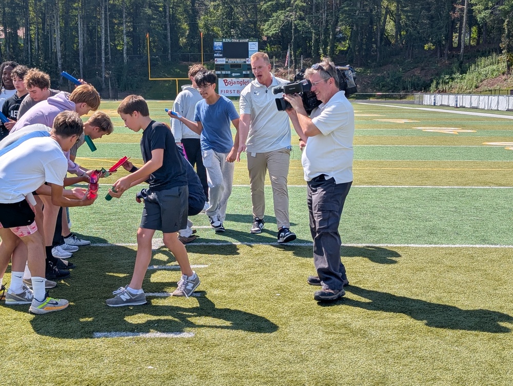 A WLOS-TV news videographer shoots footage of a teamwork activity on the the football field. Coach Greer is following a line of students holding plastic tubes.