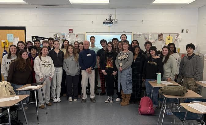 Group photo in a classroom with students gathered around Peter. 