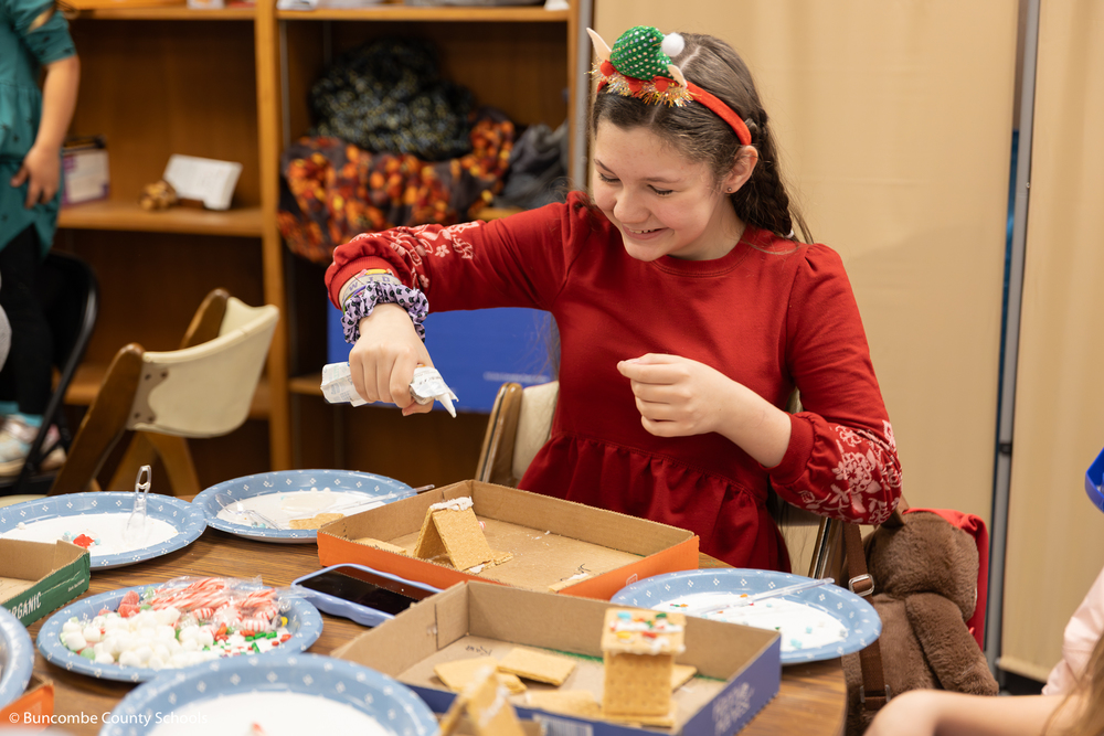 Girl wearing a red dress and elf headband putting icing on her gingerbread house to hold it together. 