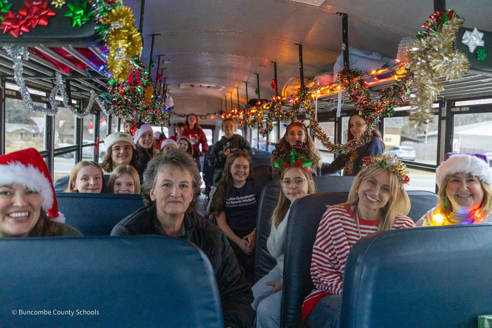 Teachers and students sit on an activity bus. Above them, they have strung tinsel, bows, holiday lights and other festive decorations.
