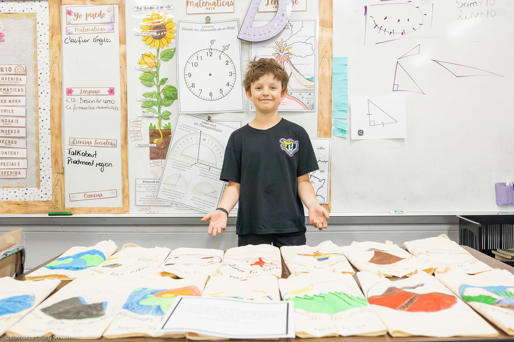 A student holds his hands out behind a table displaying hand-painted canvas bags.