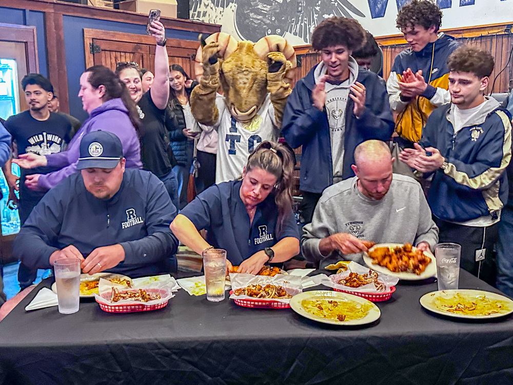 Coach Keen, Mrs. Calloway, and Coach Cook sit at a table with plates of chicken wings and baskets for bones. A crowd of fans, including the Roberson Ram mascot, is cheering behind them.