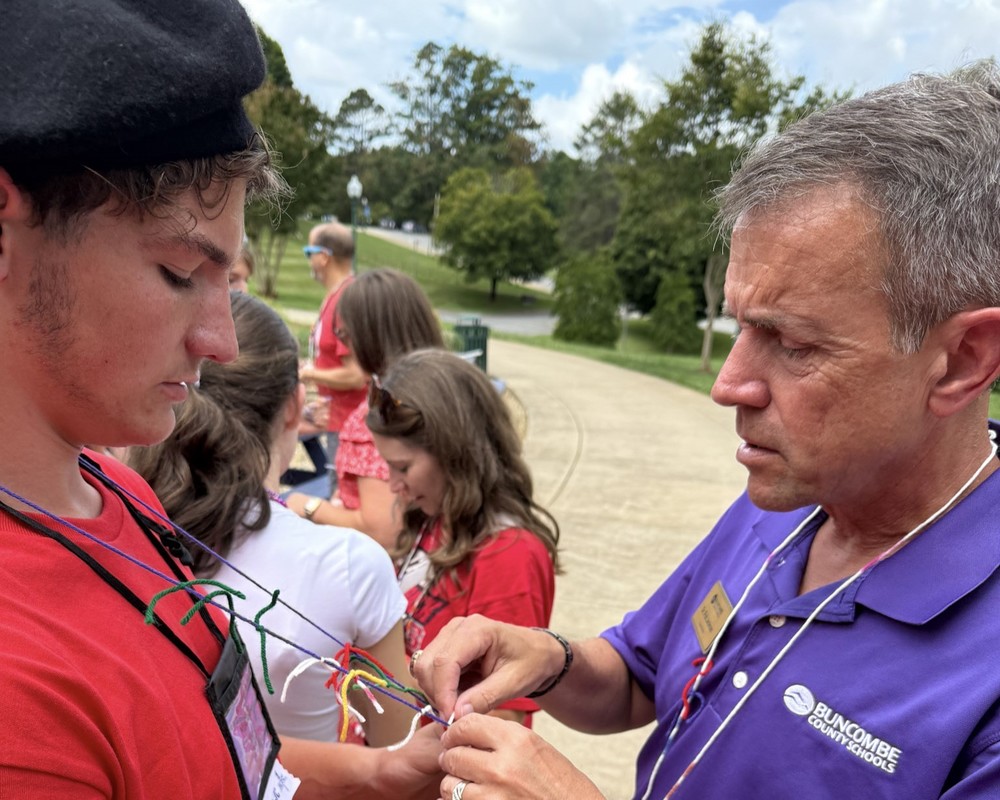 A student in a red shirt wearing a yarn necklace stands across from BCS Superintendent Dr. Rob Jackson. He is tying a ribbon around the necklace as part of a team building activity