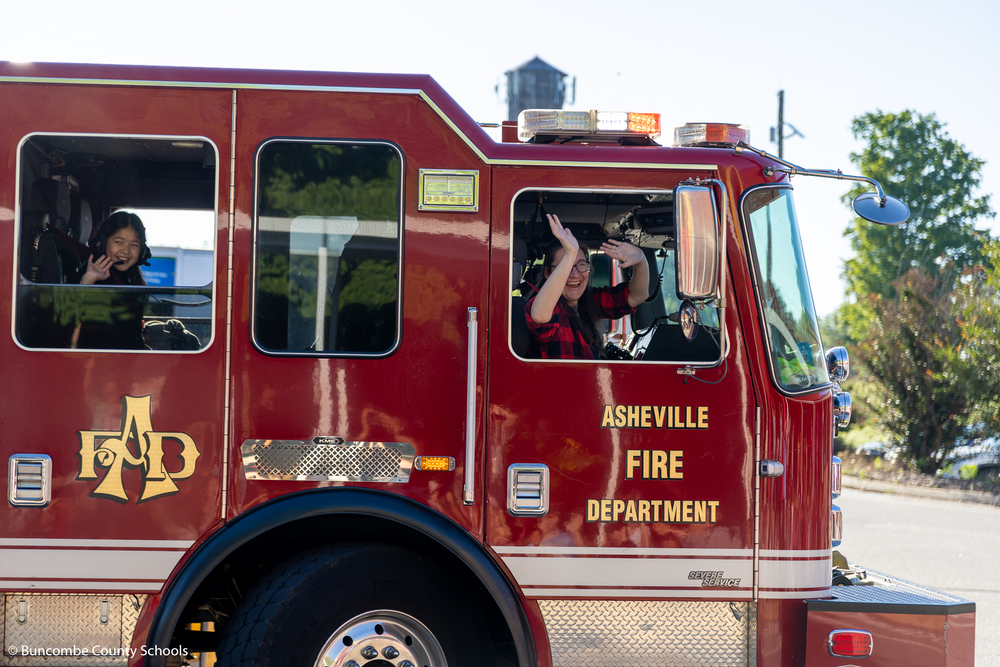 Teacher and kid waving out of the window of the firetruck as it comes around the school. 