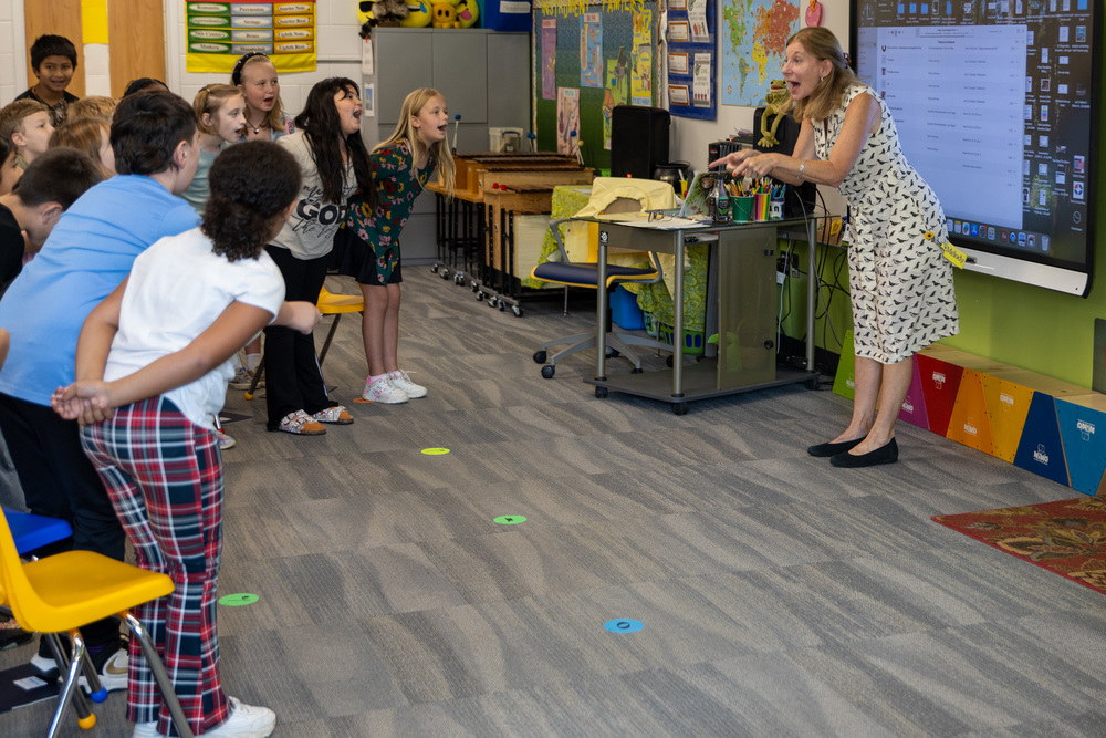 Ms. Melody stands in front of the room, facing her students who are leaning as they sing a choreographed song.