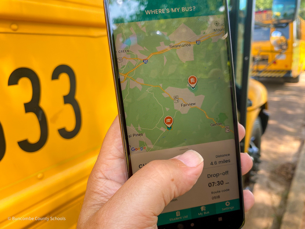 A person holds a phone in front of a school bus. The phone is displaying a map from the Edulog bus app.