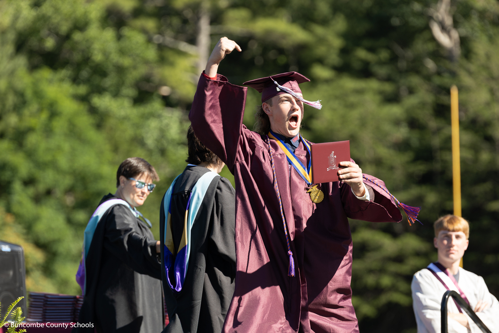 A graduate in a maroon cap and gown crosses the stgae holding up his diploma.