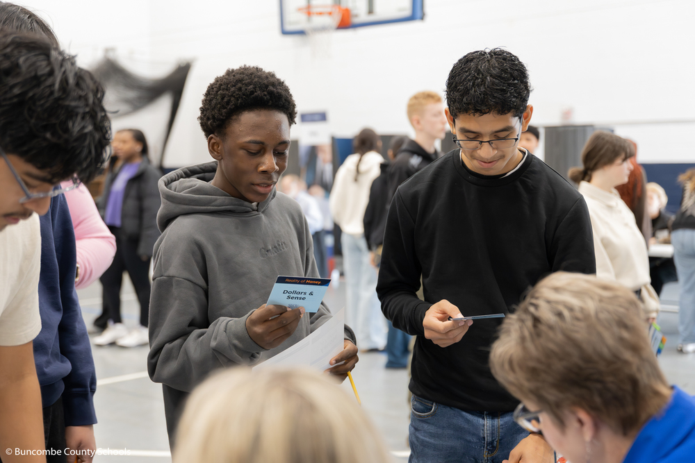 Two male students holding up a card that they drew during the Reality of Money event. They are reading what the card says.
