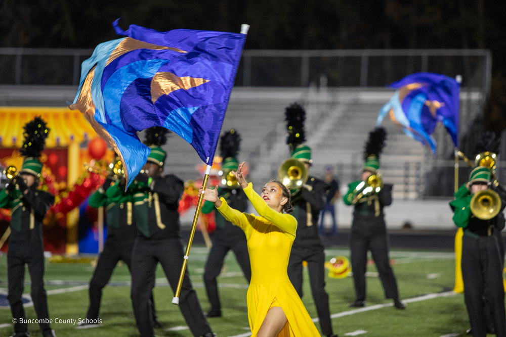 A color guard member in a yellow dress catches a blue and gold flag standing in front of the A.C. Reynolds brass section during a band performance.