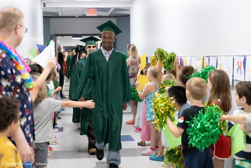 A Reynolds senior in a green cap and  down walks down a hall lined with elementary students holding pom poms