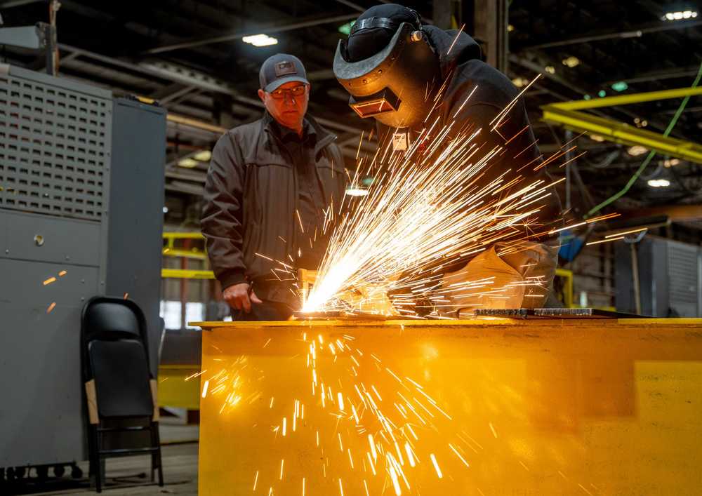 A judge observes a student wearing welding gear with sparks flying from his welder.