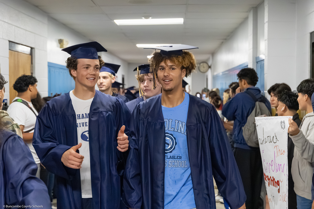 Two seniors in caps and gowns walk down a hallway lined by middle schoolers holding signs.