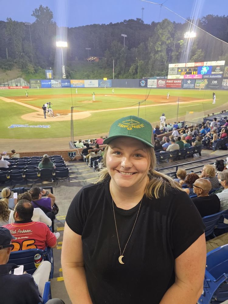 Hope Lukacena standing in the stands with the Tourist baseball game happening behind her.