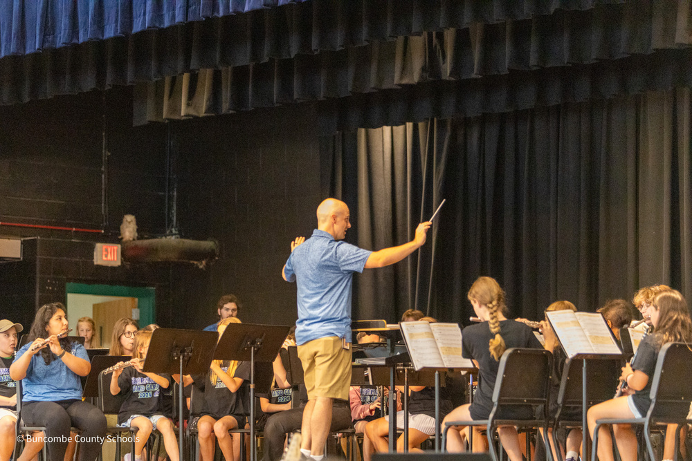 Band director Adam Stewart raises his baton as seated band students holding their instruments prepare to play a song.