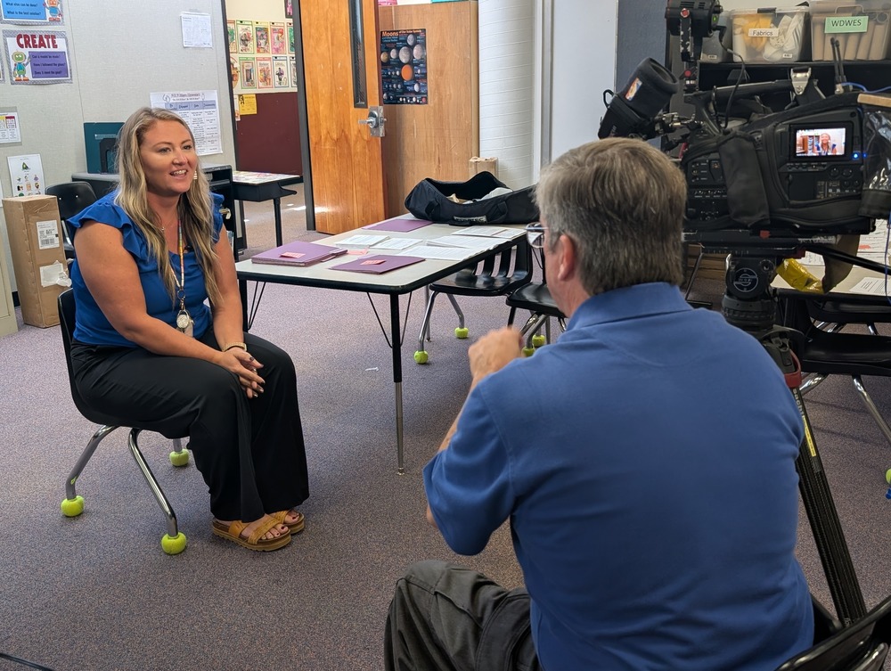 Mrs. Buchanan sits with in a classroom across from a News 13 videographer sitting next to his news camera.