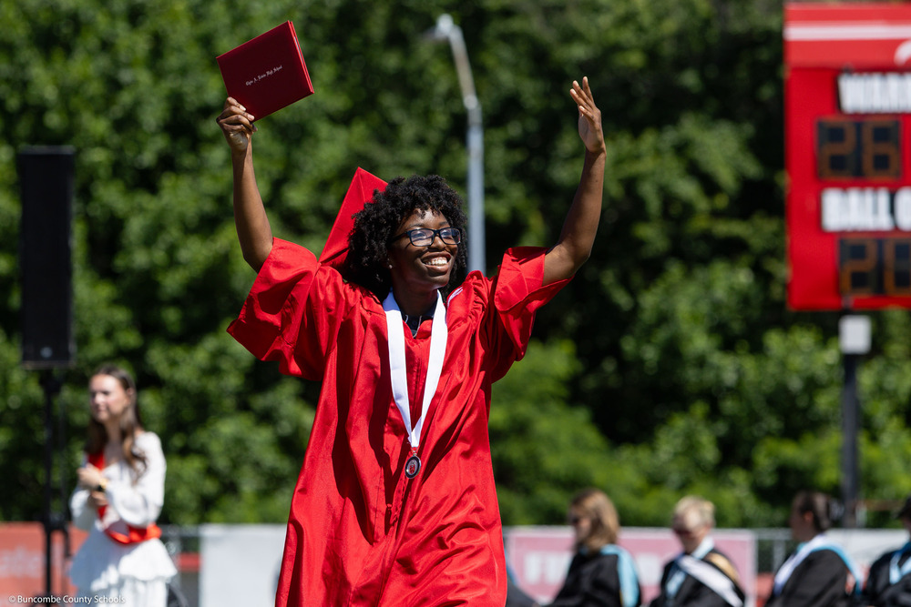 A graduate in a red cap and gown crosses the stage, smiling and waving the diploma in the air.