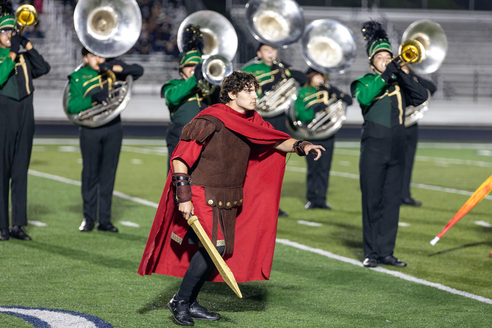 A Reynolds High band member dressed as a gladiator stands in front of students in uniform playing low brass instruments