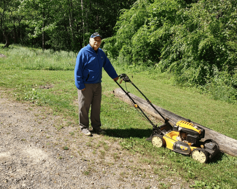 Photo of Steve pushing a yellow lawnmower at the bus stop in front of a wooden area.