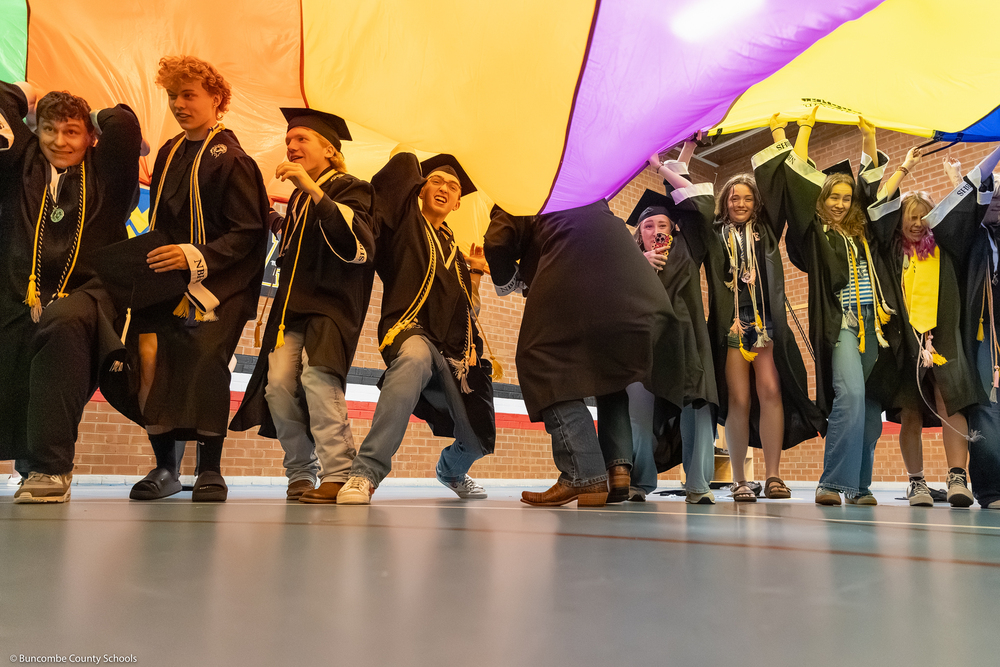 North Buncombe seniors dressed in caps and gowns get under a parachute in the school gym