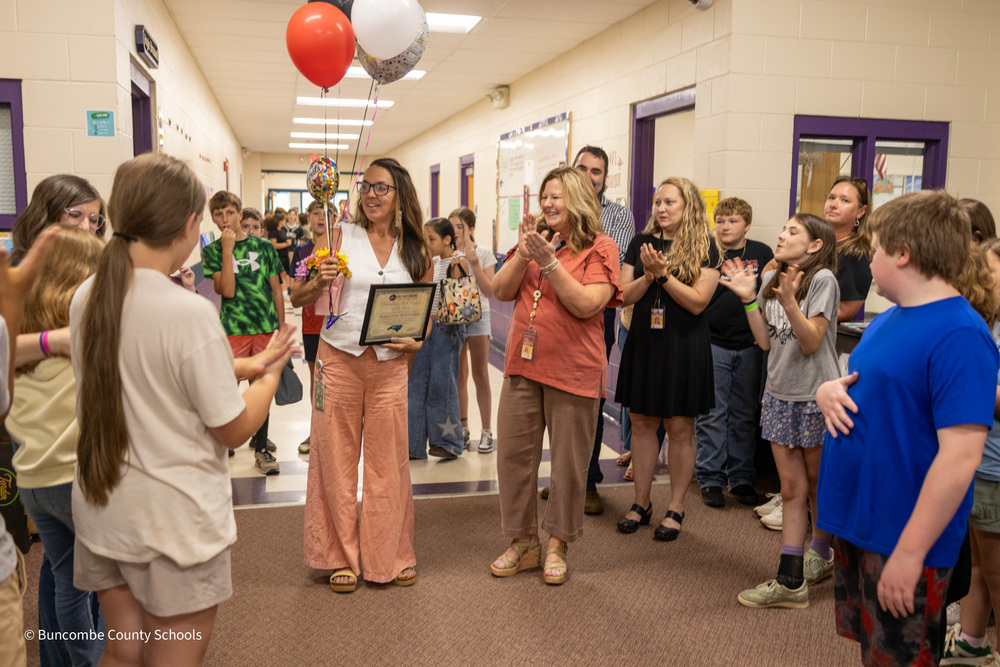Ms. Whitcher holds her plaque and balloons while  students and teachers applaud.