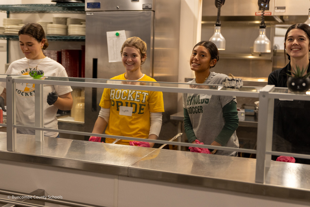 Four Reynolds students stand behind the serving line waiting to serve residents at Transformation Village