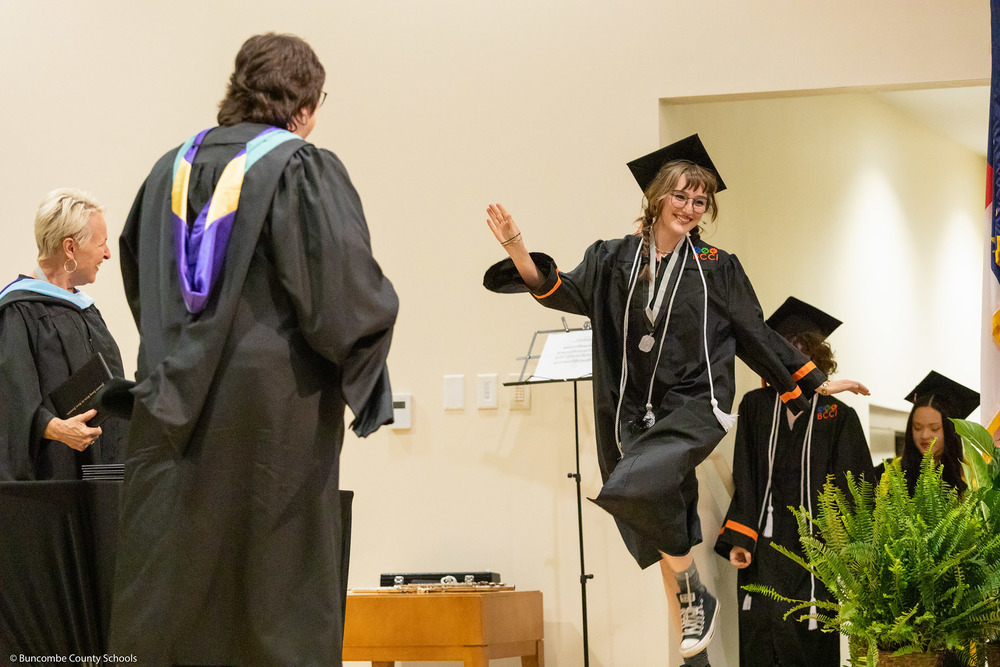 A student dressed in a cap and gown skips across the stage to receive her diploma.