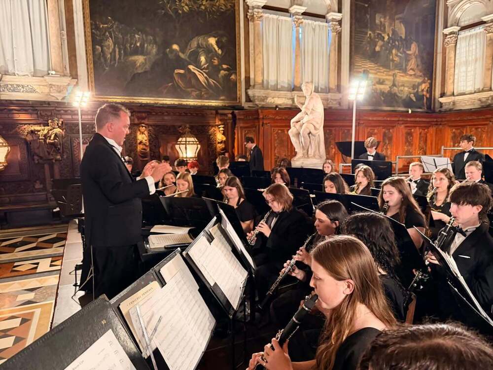 Mr. Dodson stands in an ornate stage conducting members of the band who are wearing formal wear, seated before him, playing their instruments.