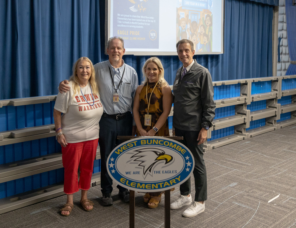 Ms. Plemmons stands next to Mr. Johnson, Ms. McCourry, and Dr. Jackson to pose for a photo. They are behind a podium with a West Buncombe Elementary sign.