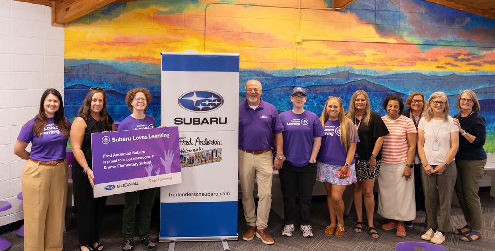 Teachers and staff at Emma Elementary pose in front of a mural in the school media center with representatives of Fred Anderson Subaru. They are standing next to a Fred Anderson Subaru "Welcome to Subieville" banner