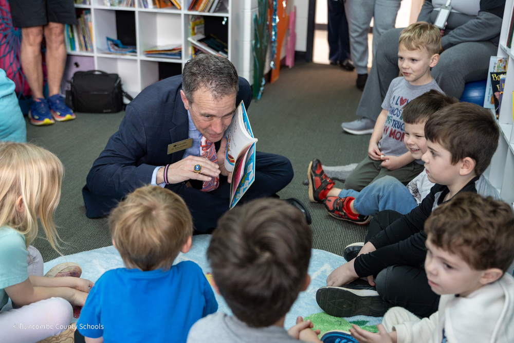 Dr. Jackson sits on the rug with preschoolers  as he holds a book. He is leaning in preparing to read the next page.