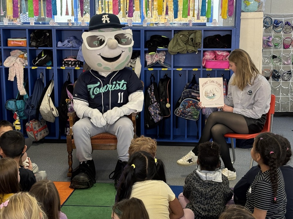 Mr. Moon the Asheville Tourist mascot sitting in a car, while his partner reads the children a book. 