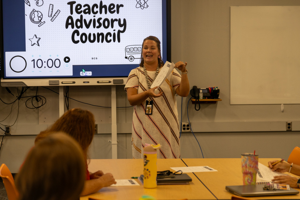 Martha Tallent stands in front of smartboard projecting "Teacher Advisory Council" slide. She is holding a paper and talking to teachers seated at tables.