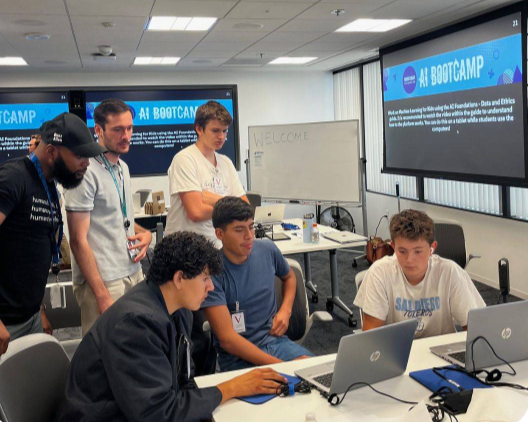 Two instructors and four students look at a laptop display. AI Bootcamp signs are displayed on big monitors around the room