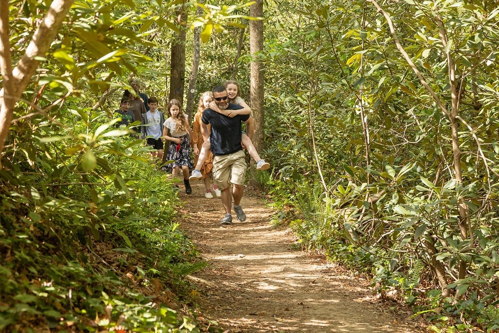 A man carries a girl on his back as a group of kids and adults walk down a trail.