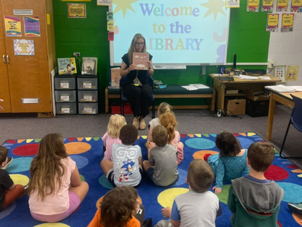 Students sit on a carpet in front of a librarian holding a book. 