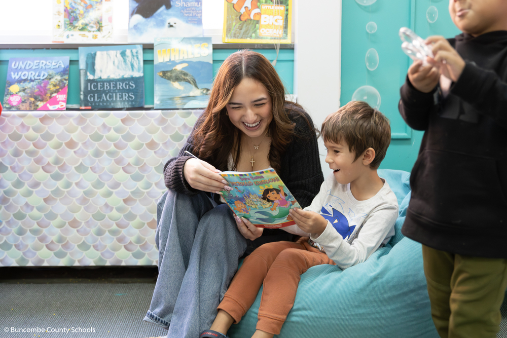 High school student and preschool student sitting on a light blue bean bag chair reading a children's book. 